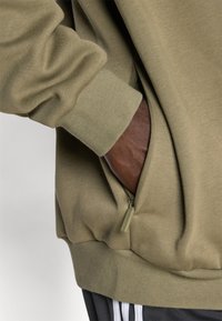 Main dans la poche d'un sweat-shirt vert olive avec un détail de fermeture éclair, montrant un poignet côtelé et un pantalon noir avec des bandes blanches.