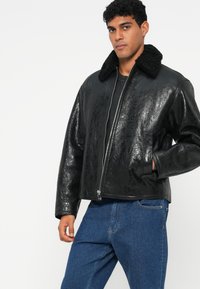 Casaco bomber de couro preto com uma textura brilhante, apresentando um colarinho felpudo e um fecho frontal, combinado com jeans azuis.