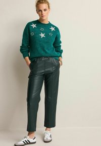 Teal gestrickter Pullover mit silbernen Sternverzierungen, kombiniert mit dunkelgrünen, taillierten Hose aus Kunstleder und weißen Sneakern mit schwarzen Streifen.