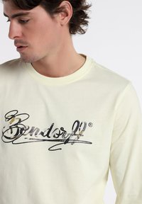 Light yellow long-sleeve shirt featuring a black and camouflage print logo. Soft fabric with a crew neckline and fitted silhouette.