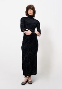 Schwarzes langärmliges Kleid mit floralem Muster, tailliertem Schnitt, hohem Kragen und transparenter Überlagerung. Kombiniert mit schwarzen, verzierten Ballerinas.