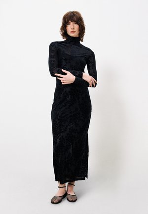 Schwarzes langärmliges Kleid mit floralem Muster, tailliertem Schnitt, hohem Kragen und transparenter Überlagerung. Kombiniert mit schwarzen, verzierten Ballerinas.