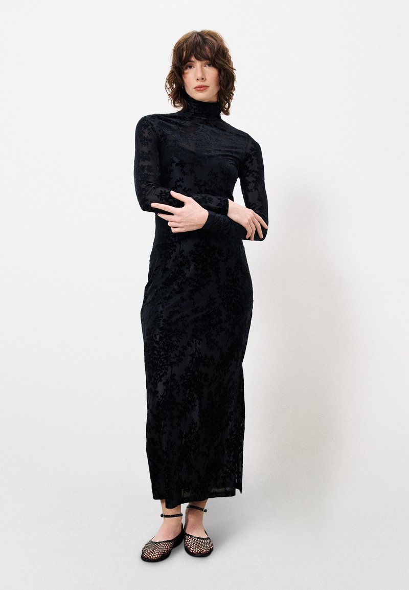Schwarzes langärmliges Kleid mit floralem Muster, tailliertem Schnitt, hohem Kragen und transparenter Überlagerung. Kombiniert mit schwarzen, verzierten Ballerinas.