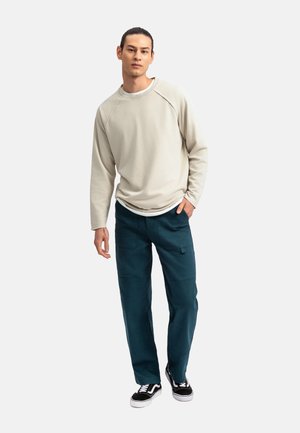 Lichtbeige sweatshirt met raglanmouwen, gecombineerd met donkerteal cargo-broek met zakken. Zwart-witte sneakers maken de outfit compleet.