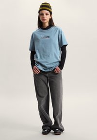 Camiseta de algodón azul claro de gran tamaño con un logo gráfico negro, usada sobre una camisa negra de manga larga, combinada con vaqueros grises holgados y zapatos negros.