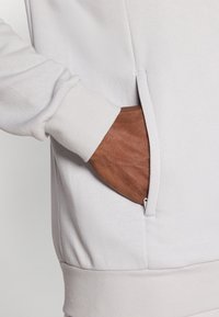 Ανοιχτό γκρι φούτερ με πλαϊνή τσέπη με αόρατο φερμουάρ, ριμπ μανσέτες και απαλό υφή στο ύφασμα.