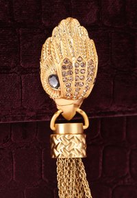 Złota ozdobna sprzączka z osadzonymi kryształami i łańcuszkowymi frędzlami na ciemnym, teksturowanym tle tkaniny.