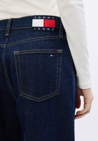 Tmavomodré džíny z denimu majú zadnú vrecko a označenie značky s červenou, bielou a modrou farbou. Šité detaily zvýrazňujú dizajn.