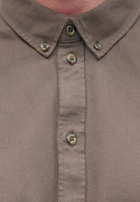 Braunes Hemd mit Knopfleiste aus einem strukturierten Stoff, versehen mit einem klassischen Kragen und fünf Knöpfen vorne, von denen zwei sichtbar sind.