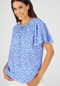 Blauwe bloemenblouse met korte vlindermouwen, voorzien van een ronde halslijn en een sleutelgatdetail. De stof heeft een gladde, lichte textuur.