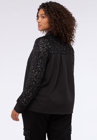Czarna bluzka z błyszczącymi ramionami i rękawami ozdobionymi cekinami, o luźnym kroju i gładkiej tkaninie, noszona z dopasowanymi spodniami.