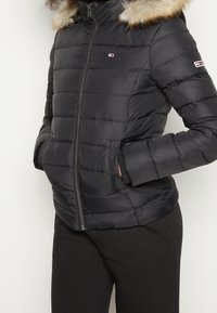 Chaqueta de plumas negra con capucha desmontable forrada de pelo, cierre frontal con cremallera y bolsillos laterales; presenta un diseño de textura acolchada horizontal y detalle del logotipo.