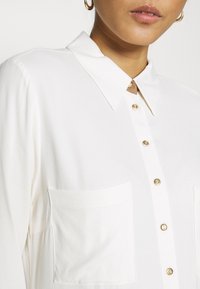 Weiße Bluse mit Knopfleiste und Kragen, goldenen Knopfakzenten und zwei vorderen Taschen. Glattes Material und taillierter Schnitt.