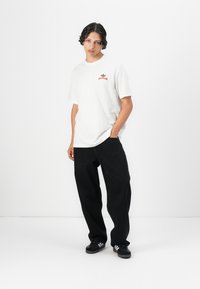 White cotton t-shirt with a red graphic logo on the chest, paired with black baggy jeans and black sneakers with white stripes.