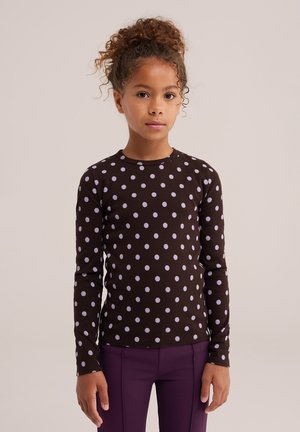 Une fille se tient debout, face à l’avant, devant un fond uni. Elle porte un haut noir à pois blancs et un pantalon violet.