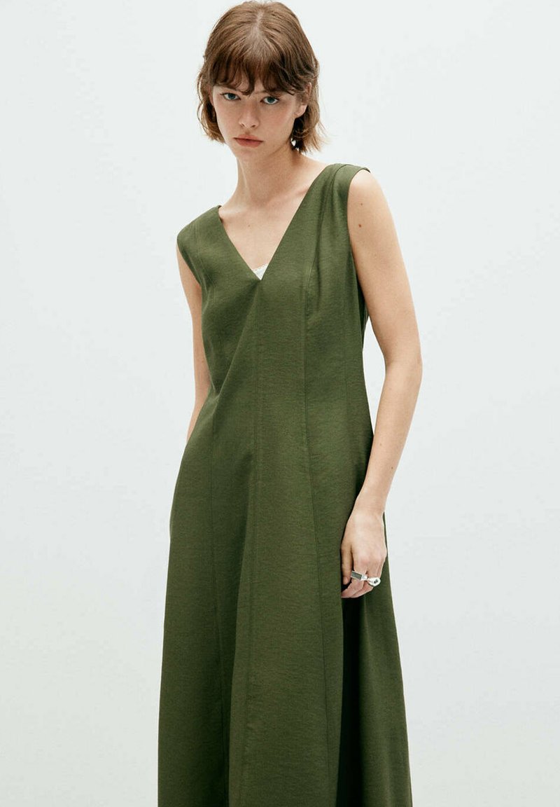 Jeune femme portant une robe sans manches vert olive avec un décolleté en V, debout devant un fond clair uni.