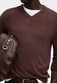 Suéter burdeos con escote en V, confeccionado en tejido suave, combinado con un bolso de cuero marrón que cuenta con un zipper dorado y un acento en forma de D.