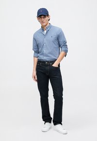 Mėlynai langeliais raštas marškiniai su mygtukais, tamsūs džinsai ir balti sportbačiai. Taip pat dėvi mėlyną kepurę su logotipu, stovi su viena ranka kišenėje.