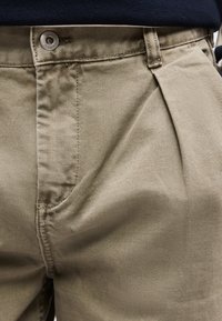 Κοντινό πλάνο μπεζ βαμβακερού παντελονιού με κούμπωμα κουμπιών, μπροστινή τσέπη και θηλιές για ζώνη, φορεμένο με σκούρο μπλε πουκάμισο.