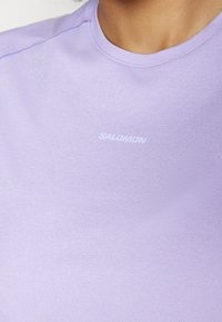 Tricou sportiv mov, dintr-un material neted, având un decolteu rotund și un logo alb "SALOMON" pe piept.