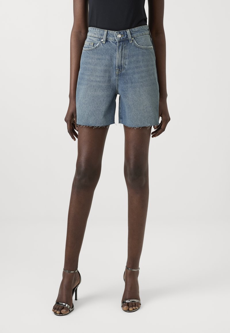 Lichtblauwe denim short met een hoge taille, gescheurde zoom, vijf zakken en een knoopsluiting, gecombineerd met zwarte schoenen met hakken.