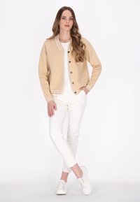 Beige gebreide cardigan met een gestreepte kraag, voorzien van zwarte knopen, gecombineerd met witte broeken en witte sneakers. Model staat voor een neutrale achtergrond.