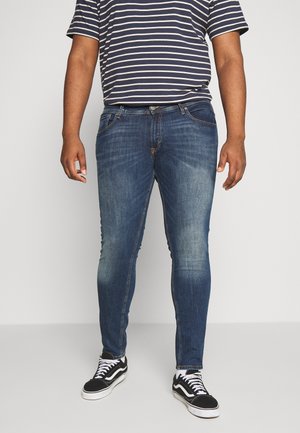 Homme portant un jean slim bleu, une chemise rayée bleu marine et blanche, et des baskets noires et blanches, debout devant un fond uni.