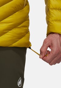 La veste isolante jaune présente un design matelassé diagonal, une taille élastique avec cordon de serrage réglable et un logo sur un pantalon vert foncé.