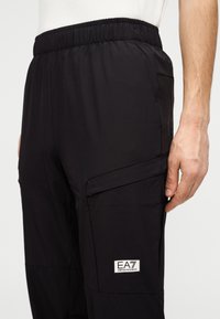 Μαύρο παντελόνι με ελαστική μέση, πλευρικές τσέπες και λογότυπο. Ελαφρύ, με λεία υφή και σχεδίαση που στενεύει προς τα κάτω για μοντέρνο ύφος.