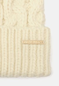 Tela de punto gruesa color crema con textura acanalada y una pequeña etiqueta rectangular de metal dorado grabada con "MICHAEL KORS."