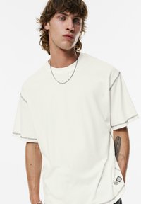 Lichtcrème oversized t-shirt met unieke dubbelgestikte korte mouwen en een klein logo-label aan de zoom. Platte gebreide halslijn en een gladde textuur.