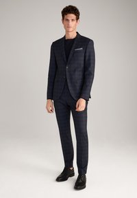 Marineblaues Karosuit mit taillierter Jacke und Hose, ausgestattet mit einem Reverskragen und einem Einzelknopfverschluss. Glattes Gewebe mit subtiler Textur. Schwarze Schuhe.