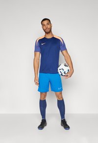 Modri nogometni šortsi Nike, temno modra majica z oranžnimi poudarki, črne kopačke in vzorčasta nogometna žoga v roki.