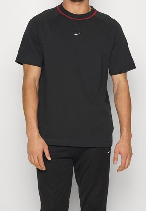Muž v černém tričku s krátkým rukávem Nike se červeným lemem kolem krku a odpovídajícími černými kalhotami Nike stojí proti jednobarevnému pozadí.