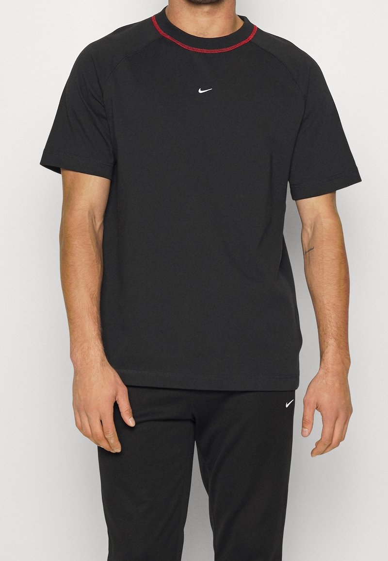 Muž v černém tričku s krátkým rukávem Nike se červeným lemem kolem krku a odpovídajícími černými kalhotami Nike stojí proti jednobarevnému pozadí.