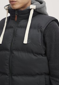 Chaleco acolchado negro con una sudadera gris debajo, que presenta un cierre con cremallera, cordones blancos y un pequeño logo en el pecho.