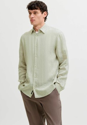 Homme portant une chemise à manches longues vert clair boutonnée et un pantalon marron, debout les mains dans les poches, regardant sur le côté.