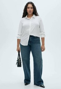 Weißes Hemd, blaue, weit geschnittene Jeans, schwarze Handtasche und spitz zulaufende schwarze Schuhe. Lockiges Haar umrahmt das Gesicht. Einfaches, lässiges Outfit.