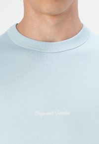 Lyseblå bomulds-sweatshirt med ribbet halsudskæring og broderet tekst "Originals Studio" i cremefarve. Glat tekstur og enkelt design.