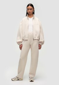 Beige zip-jakke med høj krave, parret med matchende sweatpants. Stoffet har en blød tekstur; har et logo på jakken.