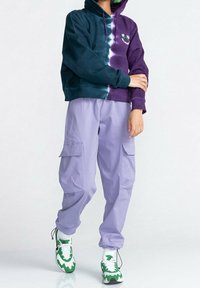 Sudadera con capucha y diseño tie-dye en verde oscuro y púrpura, con un gráfico floral. Combinada con pantalones cargo ligeros de color lavanda y zapatillas verdes.