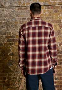 Man wearing a red and white plaid long-sleeve shirt standing against a textured brick wall, facing away from the camera.