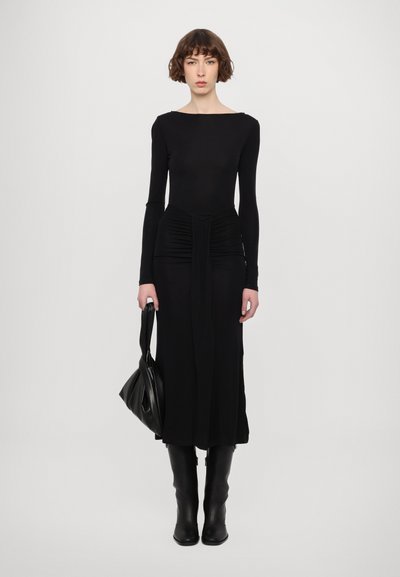 Vestido negro de manga larga con un cuerpo ajustado, detalles fruncidos en la cintura y una silueta recta. Combinado con botas negras hasta la rodilla y un bolso.