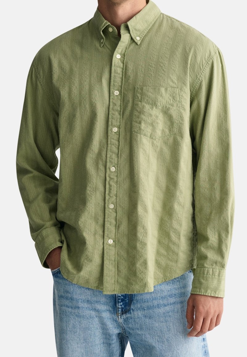 Homme portant une chemise à manches longues vert clair avec poche sur la poitrine et un jean bleu clair, debout avec une main dans la poche.