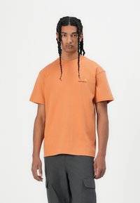 Korte mouwen oranje katoenen t-shirt met een ronde hals en een logo op de linkerborst. Model draagt donkere cargobroeken. Effen achtergrond.
