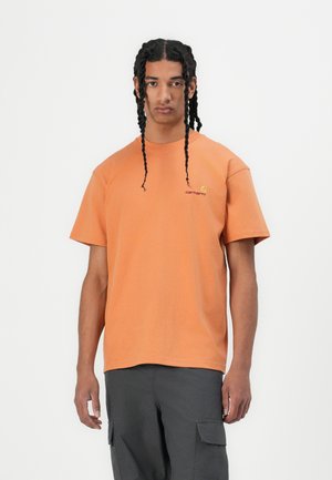 Short-sleeve orange cotton t-shirt with a crew neck and left chest logo. Model wears dark cargo pants. Plain background.