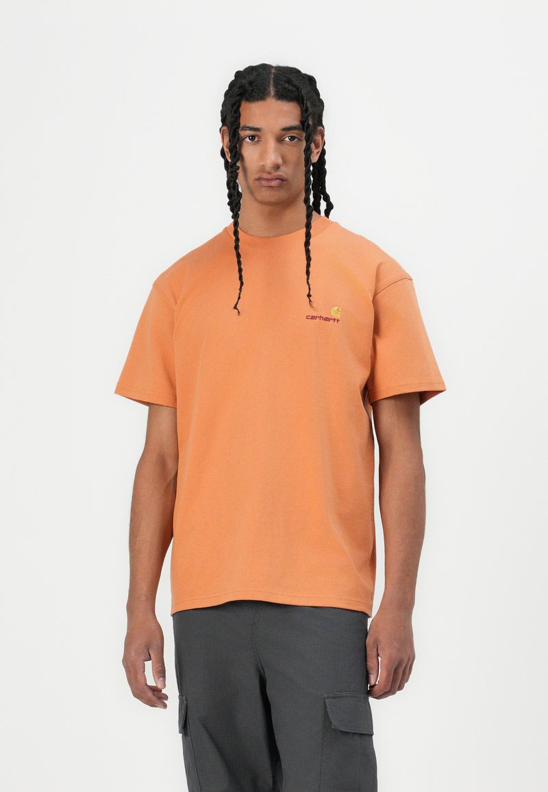 Korte mouwen oranje katoenen t-shirt met een ronde hals en een logo op de linkerborst. Model draagt donkere cargobroeken. Effen achtergrond.