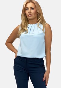Jasnoniebieski top bez rękawów z plisowaną dekoracją wokół dekoltu, zestawiony z ciemnogranatowymi spodniami z bocznymi kieszeniami i subtelnymi akcentami metalowymi.