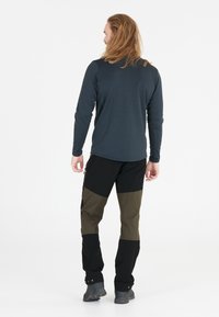 Mand med langt hår iført en mørk langærmet skjorte, tofarvede grønne og sorte udendørsbukser og mørke vandresko, stående med ryggen til.