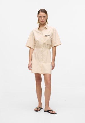 Kobieta ubrana w beżową sukienkę-koszulę z krótkim rękawem, z przodu z guzikami i kieszonką na piersi, w czarnych sandałach typu japonki, stojąca na białym tle.